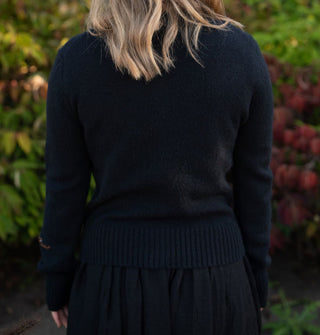 Model facing away from the camera wears a black long-sleeved sweater against a botanical backdrop