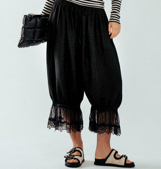 Model wears cropped black pantaloons with lace trim paired with a black and white striped long-sleeved top, puffy black clutch, and black-trimmed white woven sandals