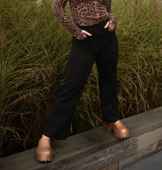 Model stands on a low stone wall in front of tall grass with hands in the pockets of black pants worn with a long-sleeved leopard print cardigan and copper clogs