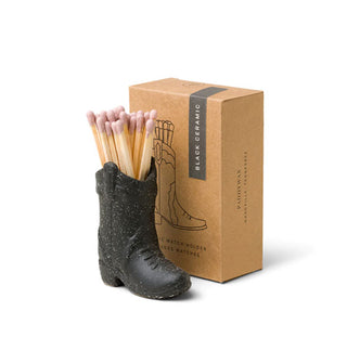 A black cowboy boot figurine holding several wooden matchsticks with light pink tips sits next to its brown box packaging