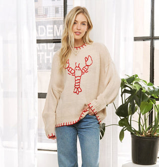 Model wears an oversized cream-colored sweater with red blanket stitching on the neck, sleeve, and bottom hems and a central red stitched lobster design
