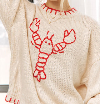 Model wears an oversized cream-colored sweater with red blanket stitching on the neckline and bottom hem and a central red stitched lobster design