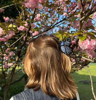 Model standing in front of a blossoming tree with pink flowers wears a blue monarch butterfly hair clip in a partially swept-back style