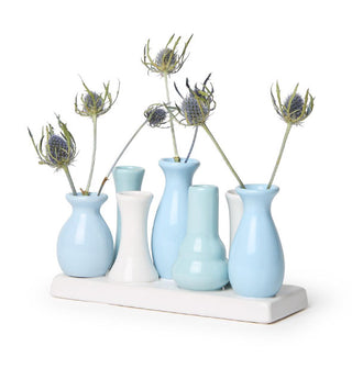 Series of small porcelain bud-sized vases in blues and whites, some holding twiggy flower buds, are clustered together on a white rectangular base