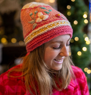 Smiling model wears a knit beanie with pink, white, and yellow striped border, pink middle with floral embroidery, and a white tip