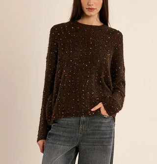 Model with one hand in pocket wears a slouchy brown sweater with multicolored fabric flecks and small sequins sewn in