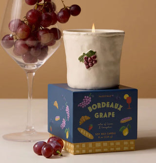 Lit Bordeaux Grape candle atop decorative gift box is staged with a wine glass and red grapes
