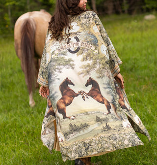 Model with back turned to the camera in a horse pasture wears a long, flowing kimono-style robe with a pastoral scene featuring two bucking horses facing one another beneath a horseshoe and banner that reads, "Born to run"