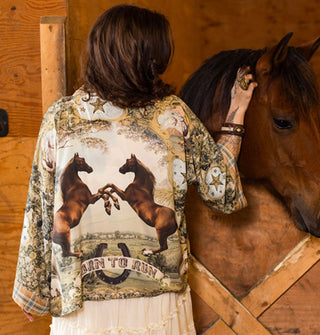 Model petting a brown horse in a stable wears an intricately designed cropped tunic with plaid cuffs and central illustration of two rearing brown horses above a banner that says, "Born to run" overtop a horseshoe