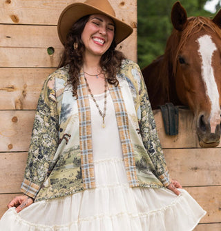 Smiling model wears a cropped open-front tunic with plaid border and densely patterned sleeves next to a stable window out of which a horse is looking