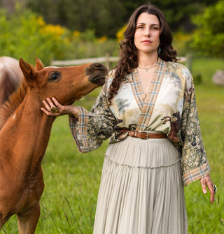 Model touching the neck of a brown foal in a green horse pasture wears an intricately patterned open-front tunic with plaid border gathered at the waist with a brown belt and paired with a white skirt