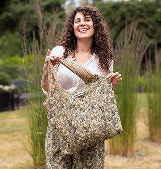 Smiling model holds a tote bag by its plaid handle that features a dense green print on its body