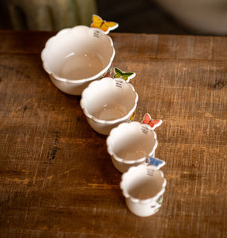 Set of four white ceramic measuring cups with butterfly handles sit in a row on a rustic wooden surface