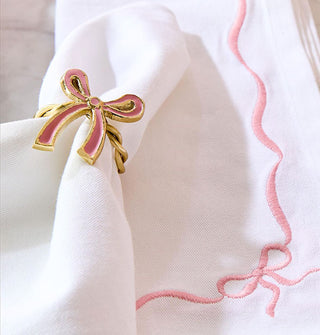 Gold napkin ring with pink enamel bow accent is placed around white linen on top of a cloth napkin with pink ribbon and bow embroidery