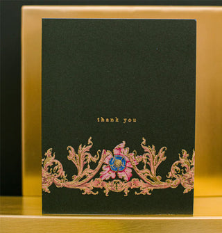 Black greeting card says, "Thank you" in small gold lettering above an ornate gold foil-accented flower and vine design
