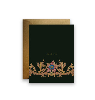 Black greeting card in front of a gold envelope says, "Thank you" in small gold lettering above an ornate gold foil-accented flower and vine design