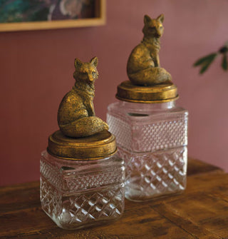 Two square embossed glass canisters with two different heights, each with a brass fox figurine lid, rest on a wooden tabletop against a dark red wall