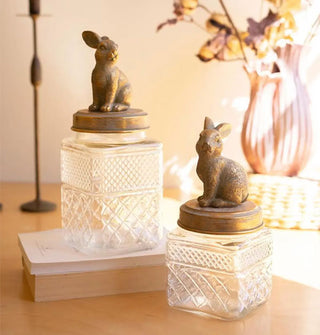 Two square crystal canisters with brass-toned rabbit figurine lids rest on a wooden tabletop staged with books, candlestick, and flower vase