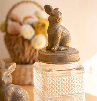 Closeup of brass-toned rabbit figurine lid atop a square embossed glass jar