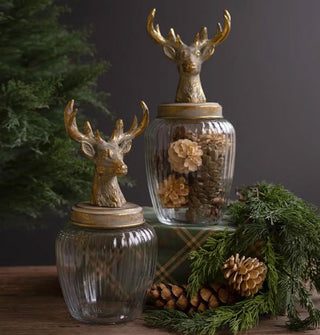 Two decorative jars with ribbed glass bodies and brass-toned antlered deer head figurine lids staged with coniferous branches and pinecones