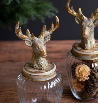 Two decorative jars with ribbed glass bodies and brass-toned antlered deer head figurine lids on a rustic wooden tabletop
