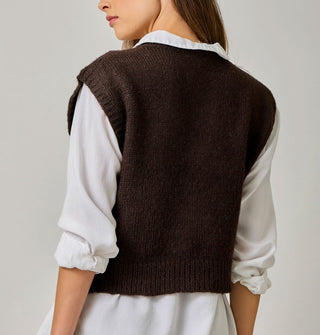 Model with back turned to the camera wears a brown sweater vest over a white collared shirt with rolled-up sleeves