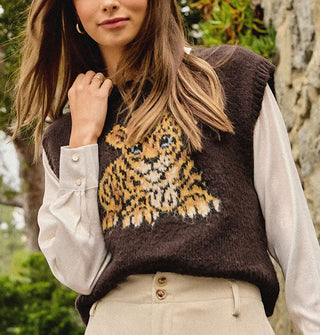 Model wears a brown sweater vest with blue-eyed leopard cub graphic over an off-white cuffed-sleeve shirt paired with tan slacks