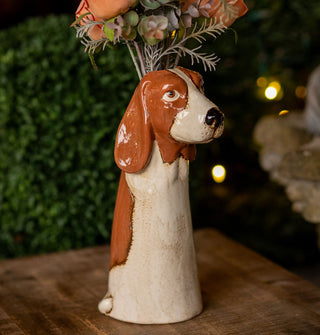 Tall dog planter/vase with brown and white patterning and long ears ears rests on a wooden tabletop against a garden backdrop with a small bouquet of flowers emerging from its top opening