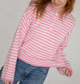 Model tugs at the bottom hem of a white and pink striped sweater with partial button placket and rolled neck