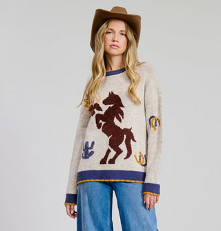 Model wears a beige sweater with brown rearing horse graphic in the center flanked by a cactus and horseshoes, more horseshoes on the left sleeve, and a blue and yellow border at the neck, cuffs, and bottom hem