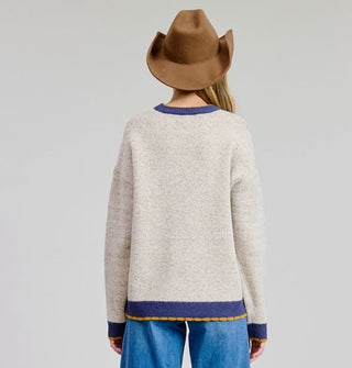 Model in cowboy hat facing away from the camera wears a beige sweater with wide blue border on the neck, cuffs, and bottom hem accented with wavy gold piping