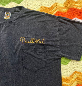 Closeup of the word, "Bullshit" embroidered in yellow script above the breast pocket of a black T-shirt