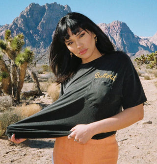 Model standing against a mountainous desert backdrop stretches out the body of a "Bullshit" embroidered black T-shirt she is wearing