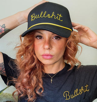 Model wears a black trucker-style hat with gold rope accent and gold embroidered script that says, "Bullshit"