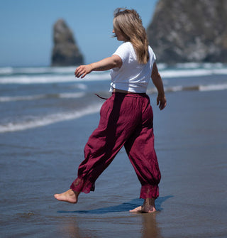 Model with arms outstretched at the shore with sea stacks in the distance wears burgundy, lace-trimmed  pantaloons with a white T-shirt
