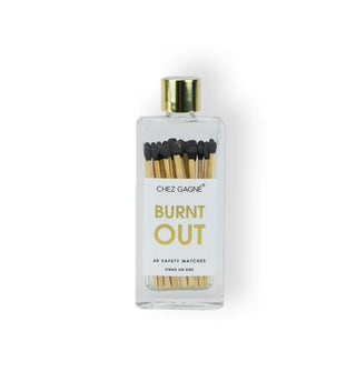 Clear rectangular glass bottle with gold cap and a label that says, “Burnt out” in gold lettering contains black-tipped wooden matchsticks