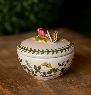 Round white ceramic lidded box with floral design, vine border, and sculptural butterfly and flower accent which serves as the lid's handle