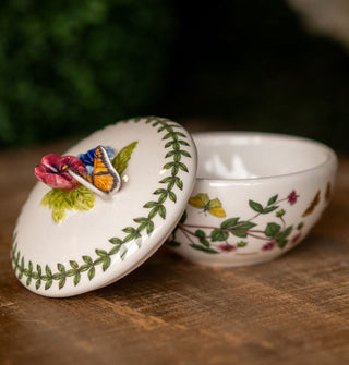 Round white ceramic lidded box with floral design, vine border, and sculptural butterfly and flower accent which serves as the lid's handle