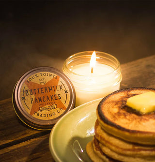 Lit Buttermilk Pancakes jar candle with lid removed is staged on a wooden surface next to a plate of pancakes with a pat of butter on top