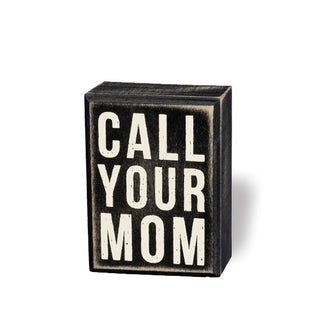Rectangular black box sign with distressed finish says, "Call your mom" in large white lettering