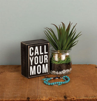Call Your Mom box sign is staged with a potted succulent and beaded bracelet on a wooden tabletop