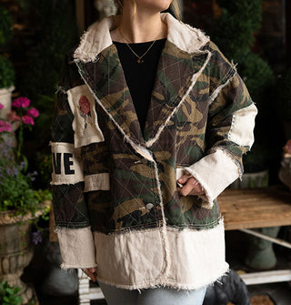 Model wears a camouflage print and white patchwork jacket with wide notched collar and raw seams against a backdrop of potted flowers