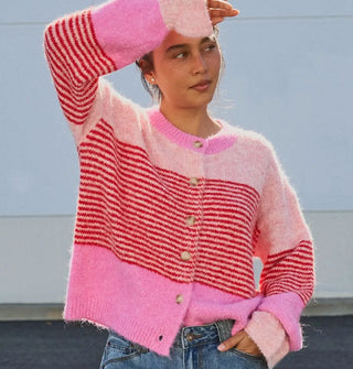 Model with one hand raised to forehead wears a button-down cardigan with gathered, voluminous sleeves and bands of light and bubblegum pink separated by a central section of dense red horizontal stripes