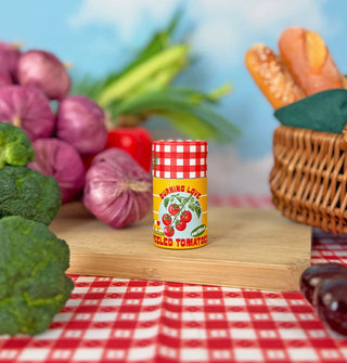 A small cylindrical container with a red and white gingham cap and label that reads, "Burning Love Italian Peeled Tomatoes" rests on a wooden cutting board surrounded by produce and a bread basket on a red and white gingham tablecloth