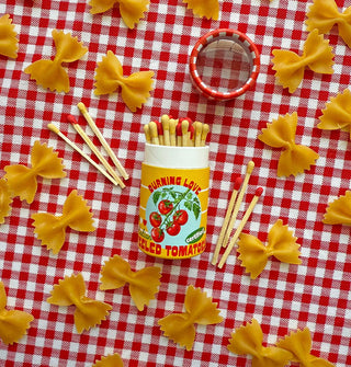 Cylindrical matchbox designed to resemble a jar of tomatoes rests on its side on a red and white gingham tablecloth scattered with bowtie pasta and some wooden matchsticks like the ones inside it, all with alternating yellow and red tips