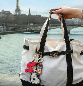 Model's hand holds the strap of a navy and white canvas tote to which are attached tomato, celestial heart, orange spritz bag charms against a Parisian skyline 