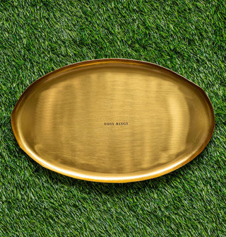 Oval-shaped gold-toned platter with shallow lip and "Rosy Rings" stamped in the center rests on green astroturf