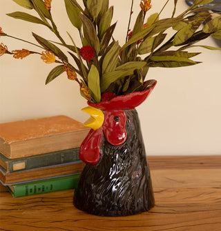 Black, red, and yellow ceramic rooster vase holds a bunch of florals on a wooden desktop staged with a stack of antique books