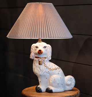 Table lamp with a pleated beige shade and a sculptural base of a hand-painted ceramic Staffordshire dog wearing a gold chain leash rests on a wooden tabletop