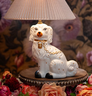 Table lamp with a pleated beige shade and a sculptural base of a hand-painted ceramic Staffordshire dog wearing a gold chain leash rests on a decorative tabletop surrounded by colorful flowers